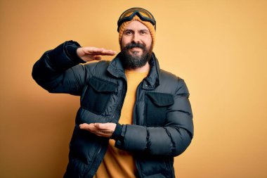 Sakallı, kar maskesi takmış, kar maskesi takmış, kayak yapan yakışıklı, kel bir kayakçı. Ellerinde büyük ve büyük beden işareti olan kayak gözlükleri var. Ölçüm sembolü. Kameraya bakıp gülümsüyor. Ölçüm konsepti.