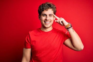 Sarışın, kıvırcık saçlı, kırmızı arka planda sıradan tişörtler giyen, parlak bir fikir ya da düşünceyle kafasını gösteren genç, yakışıklı bir adam.