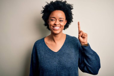 Kıvırcık saçlı, genç, Afro-Amerikan kadın. Günlük süveter giyiyor. Bir numarayı işaret ediyor ve gülümsüyor..
