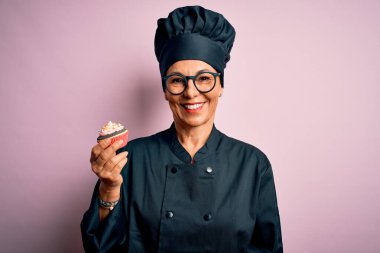 Orta yaşlı fırıncı kadın fırın üniforması giyiyor ve şapkası elinde mutlu bir yüzle duran ve gülümseyerek dişlerini gösteren lezzetli bir çikolatalı kek tutuyor.