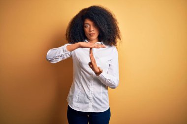 Afro saçlı, sarı arka planda dikilen güzel Afro-Amerikan zarif genç bir kadın. Elleriyle, hayal kırıklığına uğramış ve ciddi bir yüz ifadesiyle jest yapıyor.