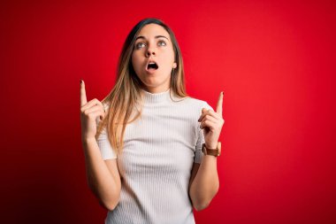 Mavi gözlü, güzel, beyaz tişörtlü, kırmızı arkaplanlı bir kadın. Şaşırmış ve şaşırmış bir şekilde yukarı bakıyor, parmaklarıyla işaret ediyor ve kollarını kaldırıyordu..