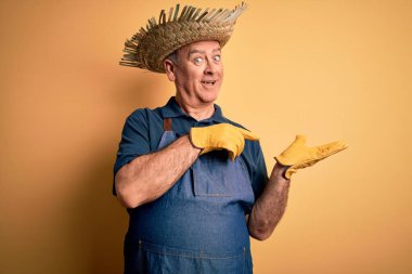 Sarı arka planda önlük ve şapka takan orta yaşlı çiftçi kameraya hayret ediyor ve gülümsüyor..