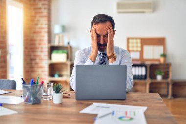 Orta yaşlı yakışıklı iş adamı ofiste dizüstü bilgisayar kullanarak oturup acı ve migrenden dolayı çaresiz ve stresli bir şekilde oturuyor. Ellerini başının üstüne koy..