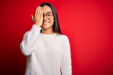 Genç, güzel Asyalı bir kadın kırmızı arka planda günlük süveter ve gözlük takıyor. Bir gözünü eliyle kapatıyor, yüzünde kendine güvenen bir gülümseme ve sürpriz bir duygu var..