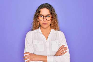 Mavi gözlü genç bir kadın mor arka planda gözlük takıyor şüpheci ve gergin, yüzünde çapraz kollu tasvip etmeyen bir ifade var. Negatif kişi.