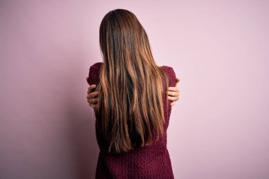 Genç, güzel bir kız, pembe arka planda rahat bir süveter giyip kendine mutlu ve pozitif bir şekilde sarılıyor. Sevgi ve özgüven.