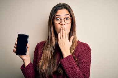 Gözlük takan genç ve güzel bir kız beyaz arka plan üzerinde akıllı telefonu gösteriyor. Ağzı kapalı, elleri utançla dolu, hata ile şok olmuş, korkunun ifadesi, sessizlik içinde korkmak, gizli bir konsept.