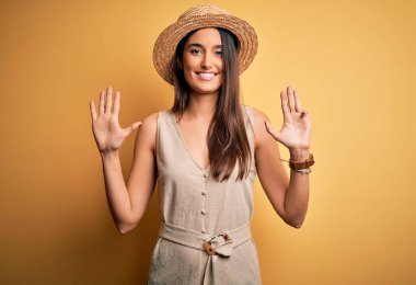 Genç ve güzel esmer kadın tatilde. Günlük elbiseler ve şapkalar giyiyor. 9 numaralı parmaklarıyla işaret ediyor ve gülümsüyor..