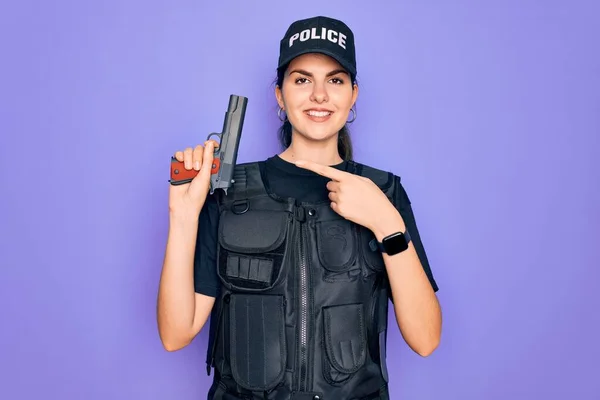 Young police woman wearing security bulletproof vest uniform and holding gun very happy pointing with hand and finger