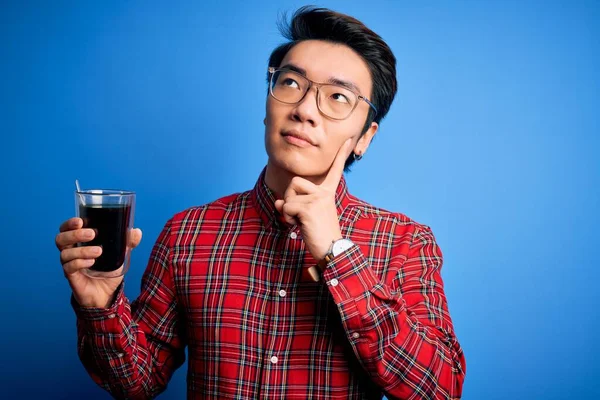 Young handsome chinese man drinking glass of coffee over isolated blue background serious face thinking about question, very confused idea
