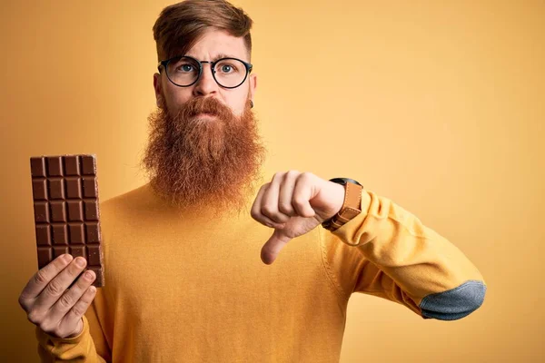 Irish redhead man with beard eating a sweet chocolate bar over yellow isolated background with angry face, negative sign showing dislike with thumbs down, rejection concept