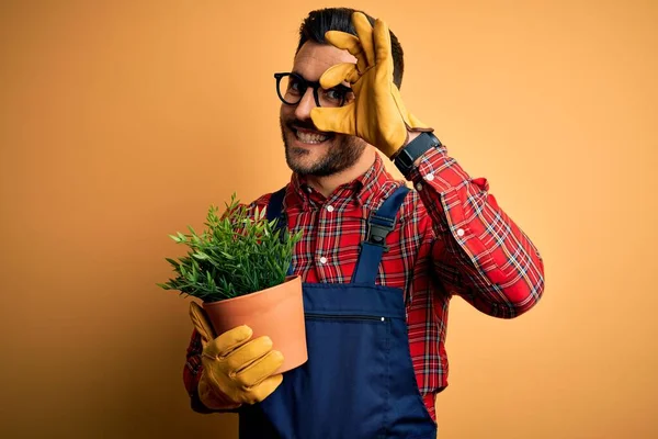 Young gardener man wearing working apron gardening plat for hobby over yellow background with happy face smiling doing ok sign with hand on eye looking through fingers