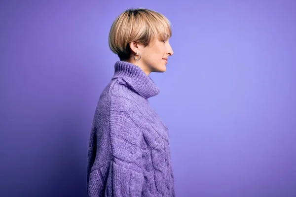 Young blonde woman with short hair wearing winter turtleneck sweater over purple background looking to side, relax profile pose with natural face and confident smile.