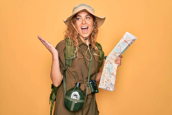 Young blonde explorer woman with blue eyes hiking wearing backpack holding city map very happy and excited, winner expression celebrating victory screaming with big smile and raised hands