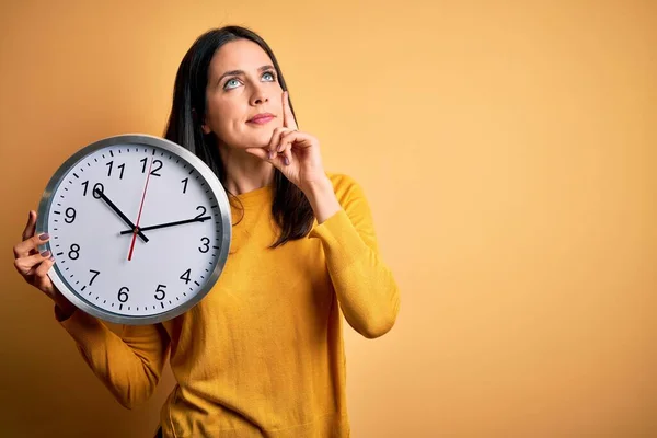 青い目の若い女性がカウントダウンをしている大きな時計を持っています黄色の背景深刻な顔を考えます質問について 非常に混乱したアイデア — ストック写真