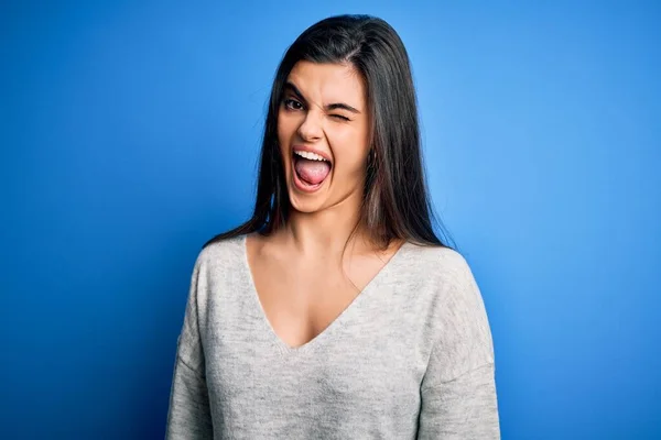 Young beautiful brunette woman wearing casual sweater standing over blue background winking looking at the camera with sexy expression, cheerful and happy face.