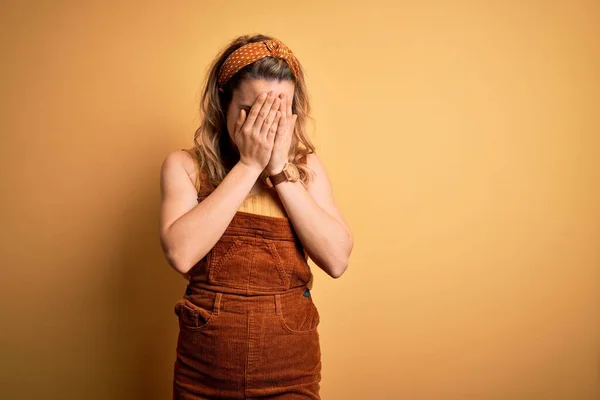 Young beautiful blonde woman wearing overalls and diadem standing over yellow background with sad expression covering face with hands while crying. Depression concept.