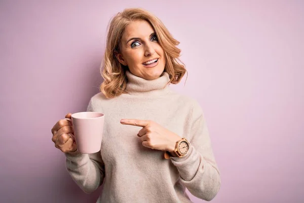 中年美丽的金发女人在与世隔绝的背景下喝着粉红色的咖啡 她非常高兴地用手和手指指指点点 — 图库照片