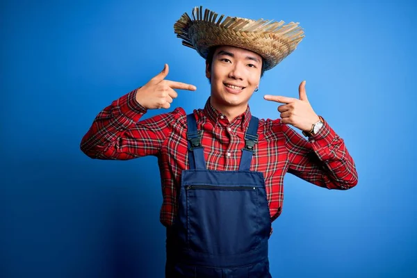 Young handsome chinese farmer man wearing apron and straw hat over blue background smiling cheerful showing and pointing with fingers teeth and mouth. Dental health concept.