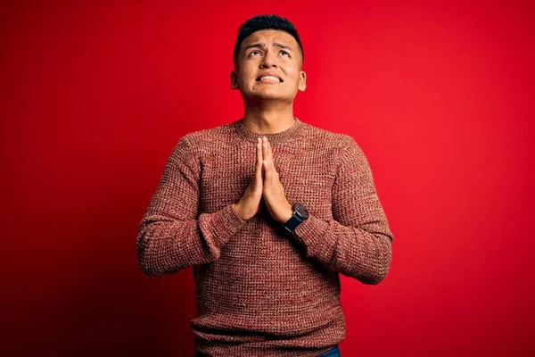 赤い背景を物乞いと手で祈る上に立ってカジュアルなセーターを着て若いハンサムなラテン人と一緒に顔に希望の表現は非常に感情的かつ心配した 物乞い — ストック写真