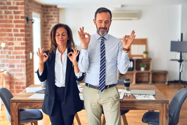 Two middle age business workers standing working together in a meeting at the office relax and smiling with eyes closed doing meditation gesture with fingers. Yoga concept.