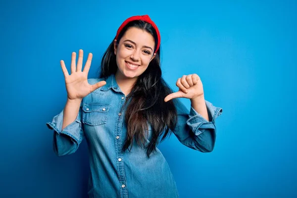 Young brunette woman wearing casual denim shirt over blue isolated background showing and pointing up with fingers number six while smiling confident and happy.