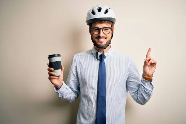 Young businessman wearing glasses and bike helmet drinking cup of coffee surprised with an idea or question pointing finger with happy face, number one