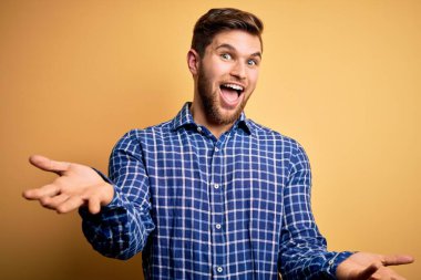 Sarışın, sakallı ve mavi gözlü genç bir işadamı sarı arka planda tişört giyiyor neşeli kollarını açıp gülümsüyor dostça karşılama, olumlu ve kendinden emin selamlar.