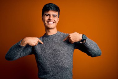 İzole edilmiş turuncu arka plan üzerinde duran genç ve yakışıklı bir adam yüzünde bir gülümsemeyle gururla ve mutlu bir şekilde kendini işaret ediyor..