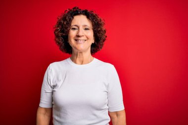 Orta yaşlı, kıvırcık saçlı güzel bir kadın izole edilmiş kırmızı arka plan üzerinde günlük tişört giyiyor ve yüzünde mutlu ve havalı bir gülümseme var. Şanslı kişi..