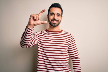 Sakallı, sıradan çizgili tişört giyen, beyaz arka planda duran, el hareketleriyle küçük boy işareti yapan, el hareketleriyle gülümseyen ve kameraya bakan genç yakışıklı bir adam. Ölçü kavramı.