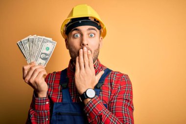 Güvenlik kaskı takan genç müteahhit elinde dolar ile sarı arka plan üstü örtülmüş ağzı, utancından ağzı açık kalmış, korkunun ifadesi, sessizlikten korkan, gizli bir konsept.