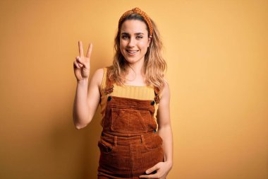 Sarı arka planda dikilip, parmaklarını zafer işareti yaparken gösteren tebessüm giymiş genç ve güzel sarışın kadın. İki numara..