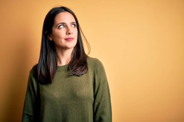 Mavi gözlü genç esmer kadın sarı arka planda yeşil süveter giyiyor gülümsüyor ve başka tarafa bakıyor..