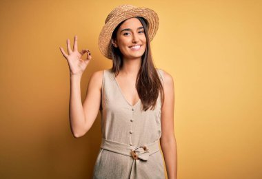 Genç ve güzel esmer kadın tatilde. Günlük elbiseler ve şapkalar giyip gülümsüyor. El ve parmaklarıyla tamam işareti yapıyor. Başarılı ifade.