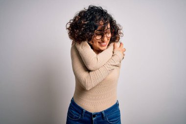 Genç ve güzel kıvırcık Arap kadın, beyaz arka planda günlük tişört ve gözlük takıyor, kendine sarılıyor, mutlu ve pozitif, kendinden emin gülümsüyor. Sevgi ve özgüven.