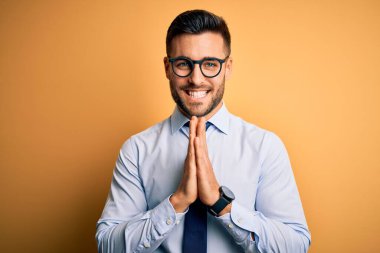 Genç yakışıklı iş adamı kravat takıyor ve gözlük takıyor sarı arka planda birlikte dua edip af diliyor kendinden emin bir şekilde gülümsüyor..
