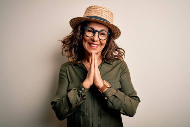 Orta yaşlı esmer kadın gözlüklü ve şapkalı. İzole edilmiş beyaz arka planda duruyor. Elleri birarada dua ediyor..