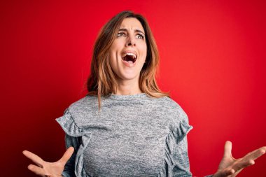 Young beautiful brunette woman wearing casual sweater standing over red background crazy and mad shouting and yelling with aggressive expression and arms raised. Frustration concept.