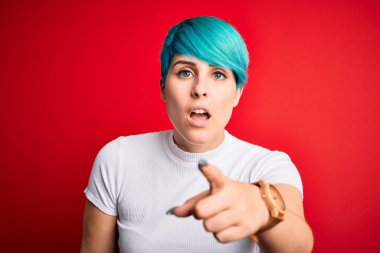 Kırmızı arka plan üzerine sıradan tişörtler giyen mavi saçlı genç ve güzel bir kadın kameraya kızgın ve sinirli olduğunu gösteriyor.