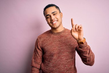 İzole edilmiş pembe arka plan üzerinde duran Brezilyalı genç bir adam bir numaralı parmağını gösterirken kendinden emin ve mutlu bir şekilde gülümsüyor..