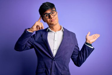 Ceket ve gözlük takan genç yakışıklı iş adamı mor arka planda kafası karışmış ve avuç içi açık bir şekilde fotokopi alanını göstermekten rahatsız olmuş ve alnını işaret ediyor. Bunu bir düşün..