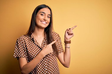 Genç, güzel esmer kadın, izole edilmiş sarı arka plan üzerinde gündelik tişört giyiyor gülümsüyor ve kameraya bakıyor iki eliyle ve parmaklarıyla yan tarafı işaret ediyor..