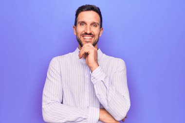 İzole edilmiş mor arka planda duran sıradan çizgili tişört giyen genç ve yakışıklı bir adam kameraya kollarını kavuşturmuş ve çenesi üzerinde kendinden emin bir şekilde gülümsüyor. Olumlu düşünüyorum..