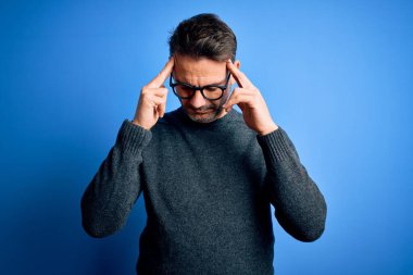 Genç, yakışıklı bir adam, günlük süveter ve gözlük takıyor. Mavi arka plan üzerinde duruyor. Başı ağrıyor çünkü stres altında. Migren ağrısı..