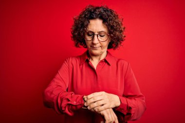 Orta yaşlı, kıvırcık saçlı, rahat gömlekli, kırmızı arka planda gözlüklü, rahat ve kendinden emin bir kadın.