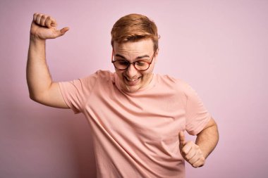 Genç, yakışıklı, günlük tişört giyen, pembe arka plan üzerinde duran mutlu ve neşeli dans eden, hareketli, rahat ve kendinden emin bir şekilde müzik dinleyen bir adam.