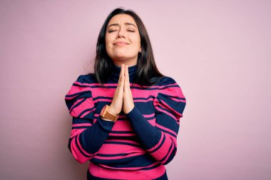 Pembe izole edilmiş arka plan üzerinde çizgili tişört giyen genç esmer bir kadın elleri bağlı yalvarıyor ve dua ediyor. Yüzünde umut ifadesi var. Çok duygusal ve endişeli. Yalvarıyorum..