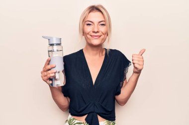 Genç, güzel sarışın kadın izole edilmiş beyaz arka planda su içiyor mutlu ve pozitif gülümsüyor, başparmağıyla mükemmel ve onay işareti yapıyor.